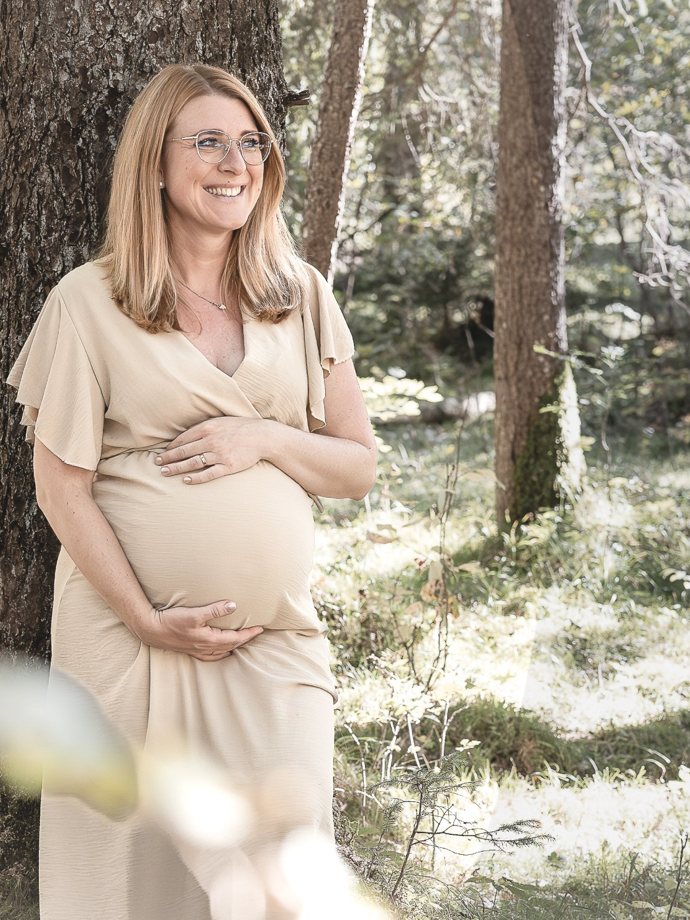 Schwangere Frau lehnt lächelnd an einem Baum im sonnigen Wald