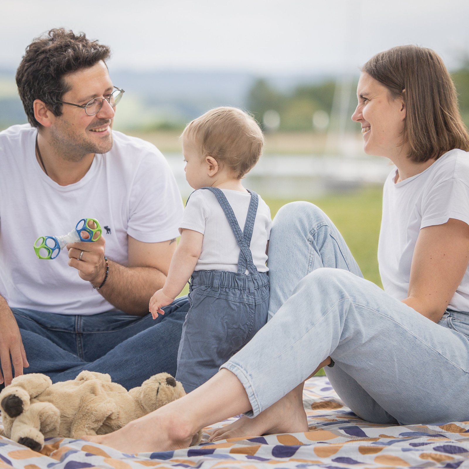 Eltern lachen beim Picknick am See, das Kleinkind steht zwischen ihnen